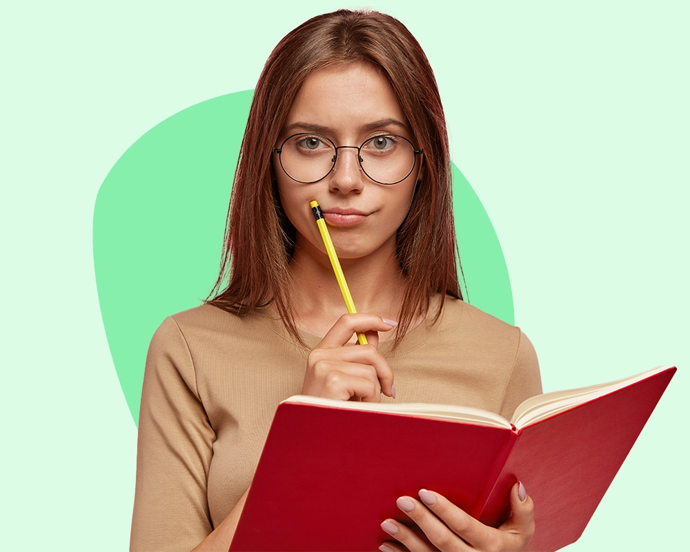 Mujer reflexiva con gafas sosteniendo un libro y un lápiz cerca de su rostro.